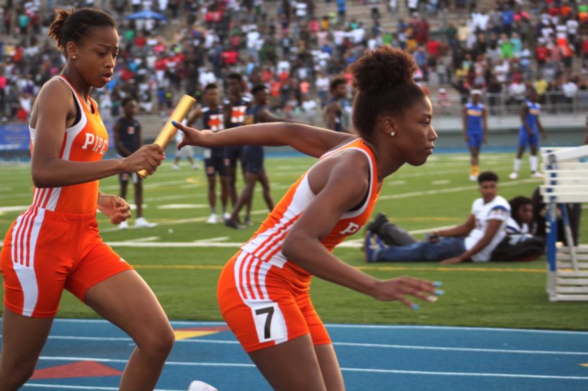 Two girls on running track, passing off relay baton.